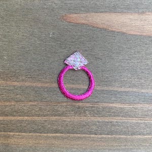 May include: A small embroidered patch in the shape of a ring. The circular band is stitched with vibrant hot pink thread, while the diamond-shaped setting at the top is filled with shimmering silver and white thread to mimic a gemstone.