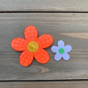 May include: Two felt flower appliqués. One large orange flower with a yellow center and stitched details. A smaller light purple flower with a green center is next to it. The flowers are on a wooden surface.