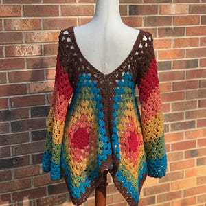 Op de afbeelding: Een kleurrijke, open geweven gehaakte top op een mannequin. De top heeft een diepe V-hals en lange mouwen. Het kleurenschema omvat bruin, rood, oranje, geel, groen en blauw, waardoor een geometrisch patroon ontstaat. Het kledingstuk is handgemaakt.