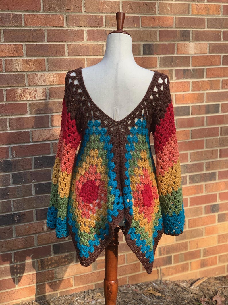 Op de afbeelding: Een kleurrijke, open geweven gehaakte top op een mannequin. De top heeft een diepe V-hals en lange mouwen. Het kleurenschema omvat bruin, rood, oranje, geel, groen en blauw, waardoor een geometrisch patroon ontstaat. Het kledingstuk is handgemaakt.