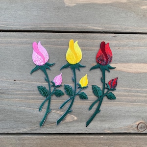 Könnte beinhalten: Drei gestickte Rosenaufnäher in Rosa, Gelb und Rot. Jeder Aufnäher zeigt eine detaillierte Rosenknospe mit grünen Blättern und Stielen. Die Aufnäher sind zum Aufnähen oder Aufbügeln auf Stoff gedacht.