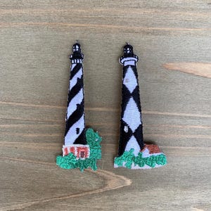 May include: Two embroidered lighthouse patches. Each patch features a black and white striped lighthouse with a red and white building at the base, surrounded by green foliage. The patches are on a wooden surface.