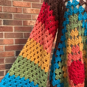 Op de afbeelding: Een handgemaakte gehaakte trui in een levendige reeks kleuren. De mouwen hebben een verloop van rood, oranje, geel en groen, terwijl het lichaam blauw en bruin bevat. Het ontwerp van de trui is een lappendeken van geometrische vormen.