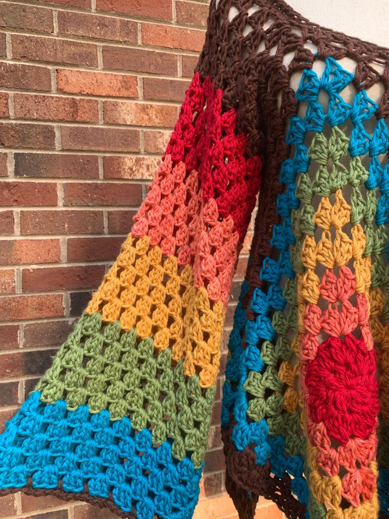 Op de afbeelding: Een handgemaakte gehaakte trui in een levendige reeks kleuren. De mouwen hebben een verloop van rood, oranje, geel en groen, terwijl het lichaam blauw en bruin bevat. Het ontwerp van de trui is een lappendeken van geometrische vormen.