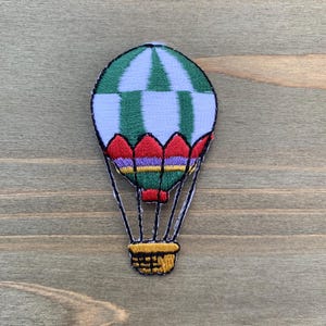 Puede incluir: Parche bordado de un globo aerostático. El globo tiene una parte superior a rayas verdes y blancas, un borde rojo y morado y una cesta amarilla. Líneas negras conectan la cesta al globo.