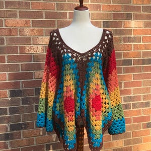 Op de afbeelding: Een kleurrijke, handgehaakte top op een mannequin. De top heeft een V-hals en lange mouwen. Het toont een geometrisch patroon in bruin, rood, geel, groen en blauw. Het kledingstuk wordt tentoongesteld tegen een bakstenen muur.
