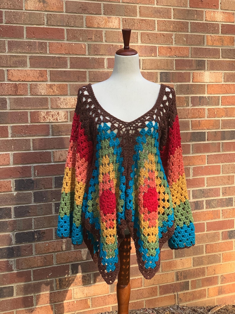 Op de afbeelding: Een kleurrijke, handgehaakte top op een mannequin. De top heeft een V-hals en lange mouwen. Het toont een geometrisch patroon in bruin, rood, geel, groen en blauw. Het kledingstuk wordt tentoongesteld tegen een bakstenen muur.