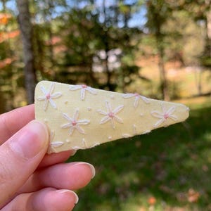 May include: A yellow fabric hair clip with white embroidered flowers and pink centers. The clip is held in a hand against a blurred green and brown background of trees and foliage. The clip is a light yellow color.