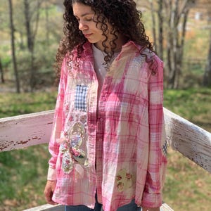 May include: Pink plaid long-sleeve shirt with patchwork details. The shirt features floral and gingham patches, and a decorative appliqué. The shirt is button-down and has a relaxed fit.