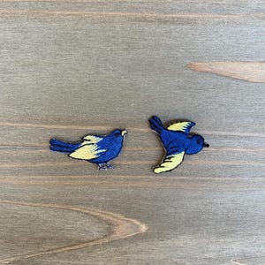 May include: Two embroidered bird patches. One bird is perched, the other in flight. Both have blue bodies and yellow wings. The patches are on a wooden surface.