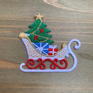 May include: An embroidered patch featuring a white sleigh with red scrollwork runners, carrying a green Christmas tree topped with a gold star and decorated with red accents, alongside blue and white gift boxes with red ribbon details.