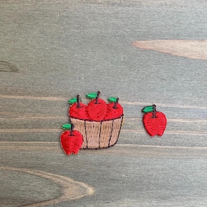 以下が含まれることがあります： 緑の葉が付いた赤いリンゴの刺繍ワッペン2点。1点は茶色の編みかごに3つの赤いリンゴが入っており、手前にもう1つ赤いリンゴが配置されている。もう1点は、緑の葉が付いた単体の赤いリンゴのワッペンである。