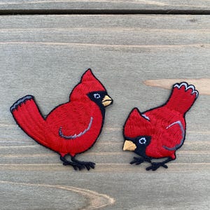Puede incluir: Dos parches bordados de pájaros cardenales rojos. Los pájaros tienen picos, ojos y patas negros. Cada pájaro tiene un cuerpo rojo con un contorno gris y un borde negro. Un pájaro está erguido y el otro está mirando hacia abajo.