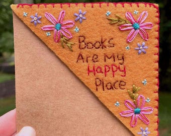 Puede incluir: Un marcapáginas de esquina triangular de fieltro color amarillo mostaza con puntada de festón roja en los bordes. Presenta flores rosas bordadas con centros turquesa, pequeñas flores azules y el texto "Books Are My Happy Place" en hilo marrón.
