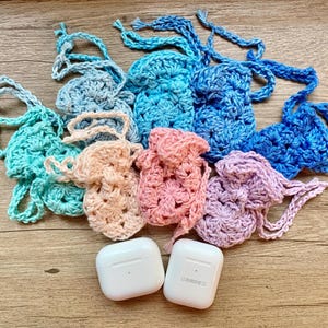 以下が含まれることがあります： 青、ターコイズ、ピンク、ラベンダーなど、さまざまな色の手作りのかぎ針編みポーチのコレクション。2つの白いワイヤレスイヤホンケースも表示されています。ポーチは小さなアイテムを収納するように設計されており、巾着クロージャーが付いています。