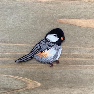 May include: Embroidered patch of a small bird with a black head, white cheeks, and gray and white wings. The bird has an orange beak and is perched on a wooden surface. The patch is detailed with various colors and textures.