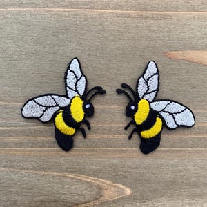 May include: Two embroidered bee patches. Each patch features a yellow and black body, white and gray wings, and black antennae. The patches are on a wooden surface.