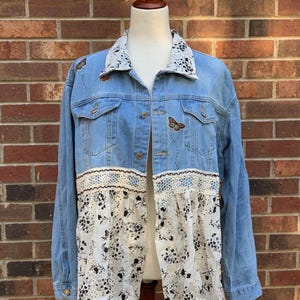 Könnte beinhalten: Hellblaue Jeansjacke mit Schmetterlingsaufnähern und einem weißen Spitzeneinsatz mit schwarzem Blumendruck. Die Jacke hat einen Spitzenkragen und ist mit kleinen weißen Pompons am Saum verziert.