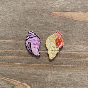 Puede incluir: Dos parches bordados de conchas marinas. Uno es de color lavanda con contornos negros. El otro es amarillo con detalles en naranja y rosa. Ambos están sobre una superficie de madera.