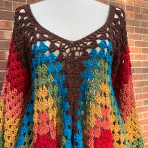 Op de afbeelding: Een kleurrijke gehaakte top met een diepe V-hals en lange mouwen. De top heeft een patchwork ontwerp in bruin, rood, oranje, geel, groen en blauw. De open weefselstructuur zorgt voor een licht en luchtig gevoel, perfect voor een bohemien stijl.