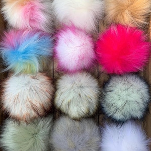 May include: A collection of twelve round faux fur pom-poms arranged in a grid. The pom-poms feature various colors including white, tan, hot pink, light blue, multi-colored pastel, grey, and tipped brown and black textures with soft fibers.