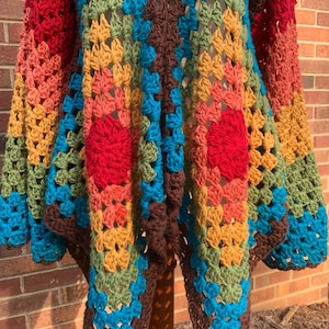 Op de afbeelding: Een handgemaakt gehaakt vest in een granny square patroon. Het open vest heeft een levendige mix van rode, oranje, gele, groene, blauwe en bruine tinten. De mouwen en het lichaam zijn versierd met een herhalend geometrisch ontwerp.