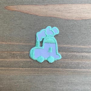 May include: An embroidered patch in the shape of a steam locomotive train. The train body is light purple with mint green wheels, a mint green roof, and a mint green smokestack emitting two puffs of light purple smoke, all outlined in mint green.