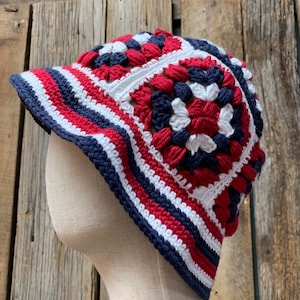 Op de afbeelding: Een handgemaakte gehaakte hoed in rood, wit en marineblauw. De hoed heeft een klep en een vierkant patroon. Het kleurenschema doet denken aan de Nederlandse vlag. De hoed wordt gepresenteerd op een mannequinhoofd tegen een rustieke houten achtergrond.