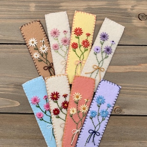 May include: A collection of handmade felt bookmarks in various colors, each embroidered with a different floral design. The bookmarks are rectangular with stitched edges and tied with a decorative string. Colors include tan, yellow, pink, red, purple, blue, and peach.
