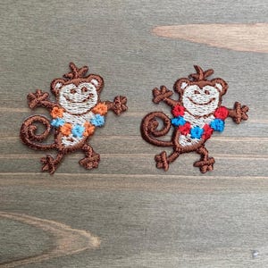 Può includere: Due toppe ricamate di una scimmia sorridente con corpo marrone, viso beige e una colorata collana di fiori. La scimmia è in posa giocosa con le braccia e le gambe tese. Le toppe sono su una superficie di legno.
