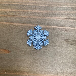 May include: A light blue embroidered snowflake patch featuring a six-pointed star design with intricate branching details and a circular center, crafted with dense thread work to create a textured, raised appearance on the surface.