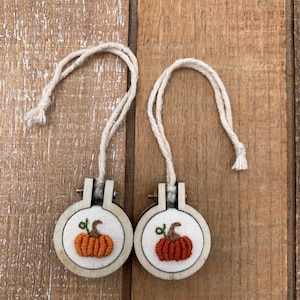 Puede incluir: Dos pequeños aros de bordado redondos con diseños de calabazas naranjas en miniatura. Cada calabaza está bordada con detalle, con un tallo y hojas verdes. Los aros son de madera clara y tienen una cuerda color crema para colgar.