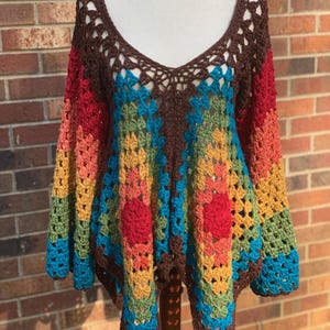 Op de afbeelding: Een kleurrijke, handgemaakte gehaakte top met een diepe V-hals. De top heeft een patchwork-ontwerp met een bruine halslijn en mouwen, die overgaan in levendige strepen van rood, oranje, geel, groen en blauw. De open weving voegt een delicate touch toe.