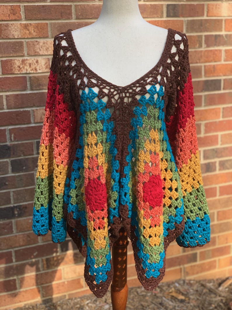 Op de afbeelding: Een kleurrijke, handgemaakte gehaakte top met een diepe V-hals. De top heeft een patchwork-ontwerp met een bruine halslijn en mouwen, die overgaan in levendige strepen van rood, oranje, geel, groen en blauw. De open weving voegt een delicate touch toe.