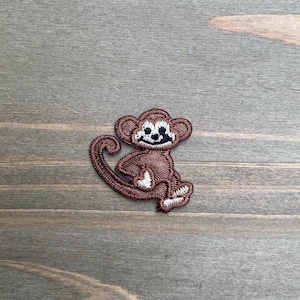May include: Embroidered patch featuring a smiling monkey in shades of brown, white, and black. The monkey is sitting with its tail curled. The patch is designed for application on clothing or accessories.