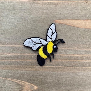 May include: An embroidered iron-on patch shaped like a bumblebee. The bee features a black head, a yellow and black striped abdomen, and two white wings with black outlines. The design includes two black antennae and small black legs.
