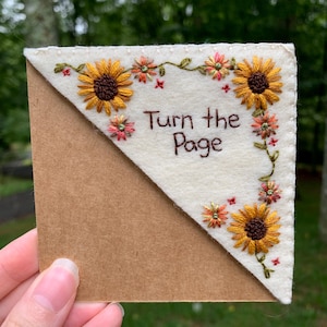 May include: Embroidered corner bookmark featuring sunflowers and the text "Turn the Page." The bookmark has a cream-colored felt top with floral embroidery and a brown paper backing. The design includes yellow, orange, and green thread.
