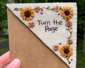 Puede incluir: Marcapáginas de esquina bordado con girasoles y el texto "Turn the Page". El marcapáginas tiene una parte superior de fieltro color crema con bordados florales y un reverso de papel marrón. El diseño incluye hilo amarillo, naranja y verde.