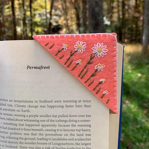 May include: A triangular coral-colored felt corner bookmark featuring hand-embroidered white daisy flowers with yellow centers and green stems. The edges of the bookmark are finished with a white blanket stitch, and it is placed on a book page.