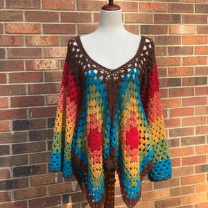 Op de afbeelding: Een kleurrijke, handgemaakte gehaakte top met een V-hals. De top heeft een levendig patroon van rood, oranje, geel, groen en blauw, met bruine afwerking en lange mouwen. Het kledingstuk wordt op een mannequin gepresenteerd tegen een bakstenen muur.