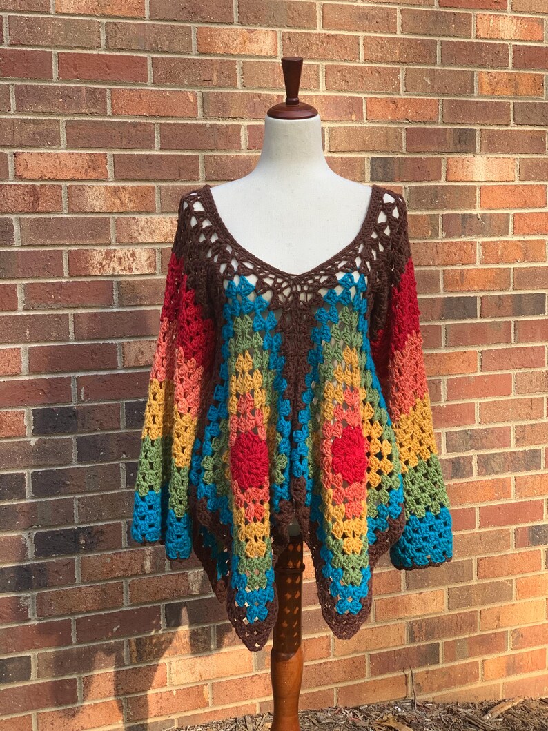 Op de afbeelding: Een kleurrijke, handgemaakte gehaakte top met een V-hals. De top heeft een levendig patroon van rood, oranje, geel, groen en blauw, met bruine afwerking en lange mouwen. Het kledingstuk wordt op een mannequin gepresenteerd tegen een bakstenen muur.