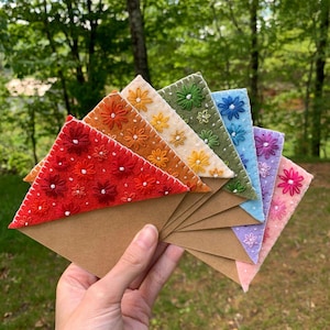 Könnte beinhalten: Eine Hand hält einen Fächer aus bunten Filzdreiecken mit gestickten Blumenmustern. Jedes Dreieck hat eine andere Farbe, darunter Rot, Orange, Gelb, Grün, Blau, Lila und Rosa, und ist an einem braunen Papierumschlag befestigt.