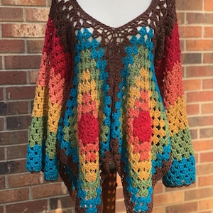 Op de afbeelding: Een kleurrijke, handgemaakte gehaakte top met een V-hals. De top heeft een bruine halslijn en mouwen, met een levendig patroon van rood, oranje, geel, groen en blauw. Twee rode bloemachtige accenten zijn zichtbaar aan de voorkant.