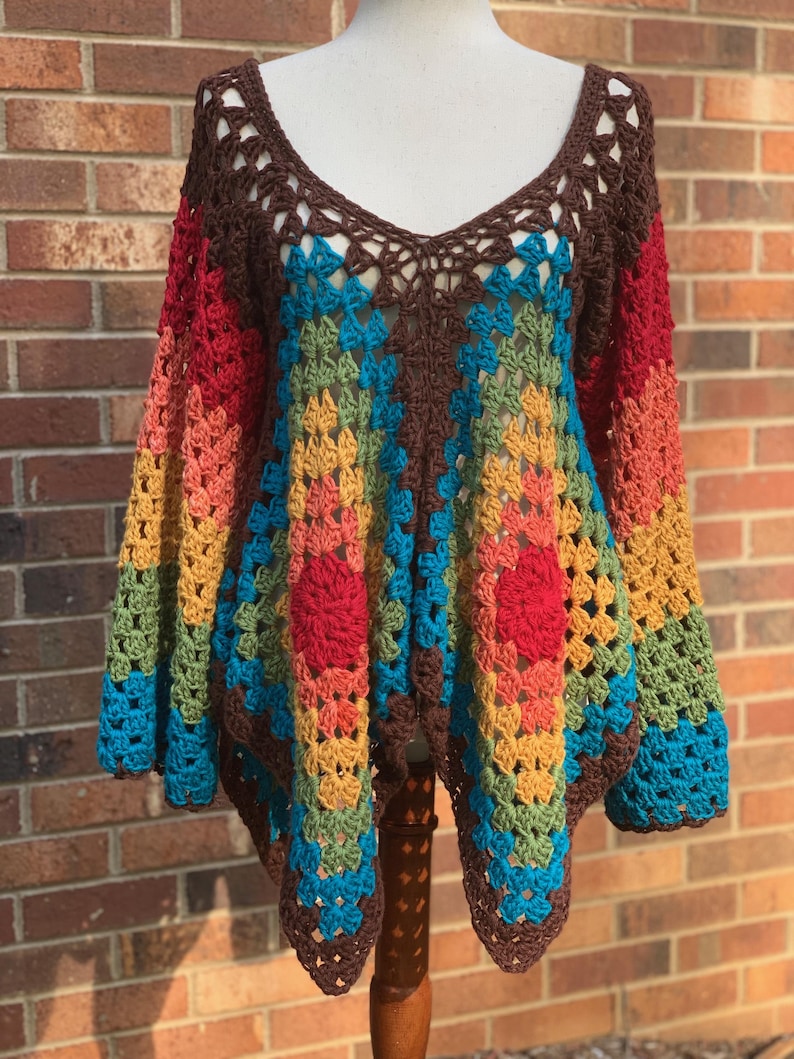 Op de afbeelding: Een kleurrijke, handgemaakte gehaakte top met een V-hals. De top heeft een bruine halslijn en mouwen, met een levendig patroon van rood, oranje, geel, groen en blauw. Twee rode bloemachtige accenten zijn zichtbaar aan de voorkant.