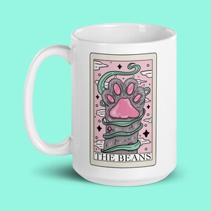 May include: White ceramic mug featuring a cartoon illustration of a cat's paw wrapped in vines. The illustration is set against a pink and white background with the text "THE BEANS" at the bottom.