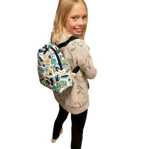 May include: A young person wearing a grey sweater with a unicorn print and black leggings is wearing a white backpack with a colorful dinosaur print.