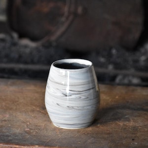 May include: A handmade ceramic tumbler with a marbled gray and white pattern. The small, rounded vessel has a dark interior and sits on a wooden surface. The background is blurred, suggesting a rustic setting.