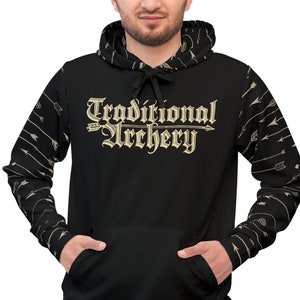 以下が含まれることがあります： ヴィンテージフォントで「Traditional Archery」と書かれた黒いパーカー。袖には矢の模様があります。パーカーにはフロントポケットとフードが付いています。その人は濃い色のズボンを履いています。