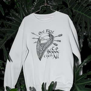 以下が含まれることがあります： 矢が刺さった盾が描かれた黒いグラフィックデザインの白いスウェットシャツ。テキストは「One Ring To Shoot Them All」です。デザインは様式化されたイラストスタイルです。