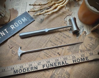 May include: A silver-coloured metal reflex hammer with a cylindrical head and a smooth handle, positioned above a silver-coloured metal surgical instrument with a T-shaped handle marked with the word FREEMAN, resting near a wooden ruler.